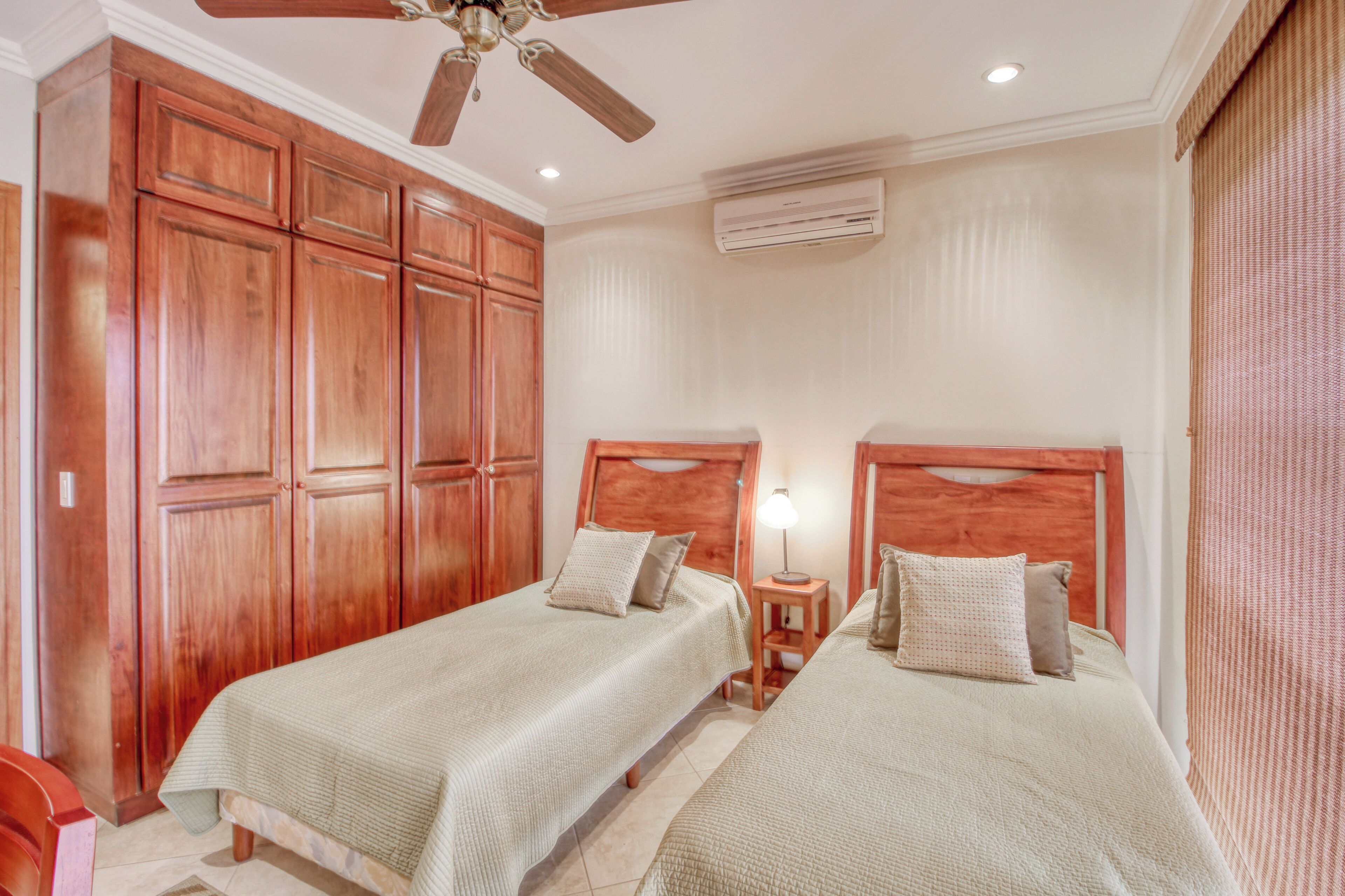 A bedroom with two beds, wooden wardrobes, a ceiling fan, an air conditioning unit, and a bedside lamp.