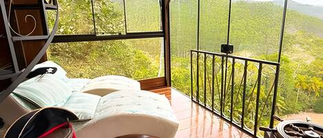 A bedroom with a bed, a wooden floor, a window with a view of greenery, and a balcony with a railing.