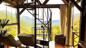 A room with a view of green hills, a chair, a table, and a small cabinet.