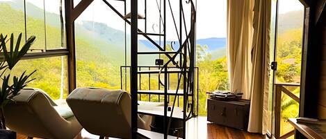 A room with a view of green hills, a chair, a table, and a small cabinet.