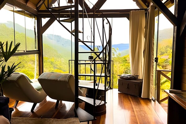 A room with a view of green hills, a chair, a table, and a small cabinet.