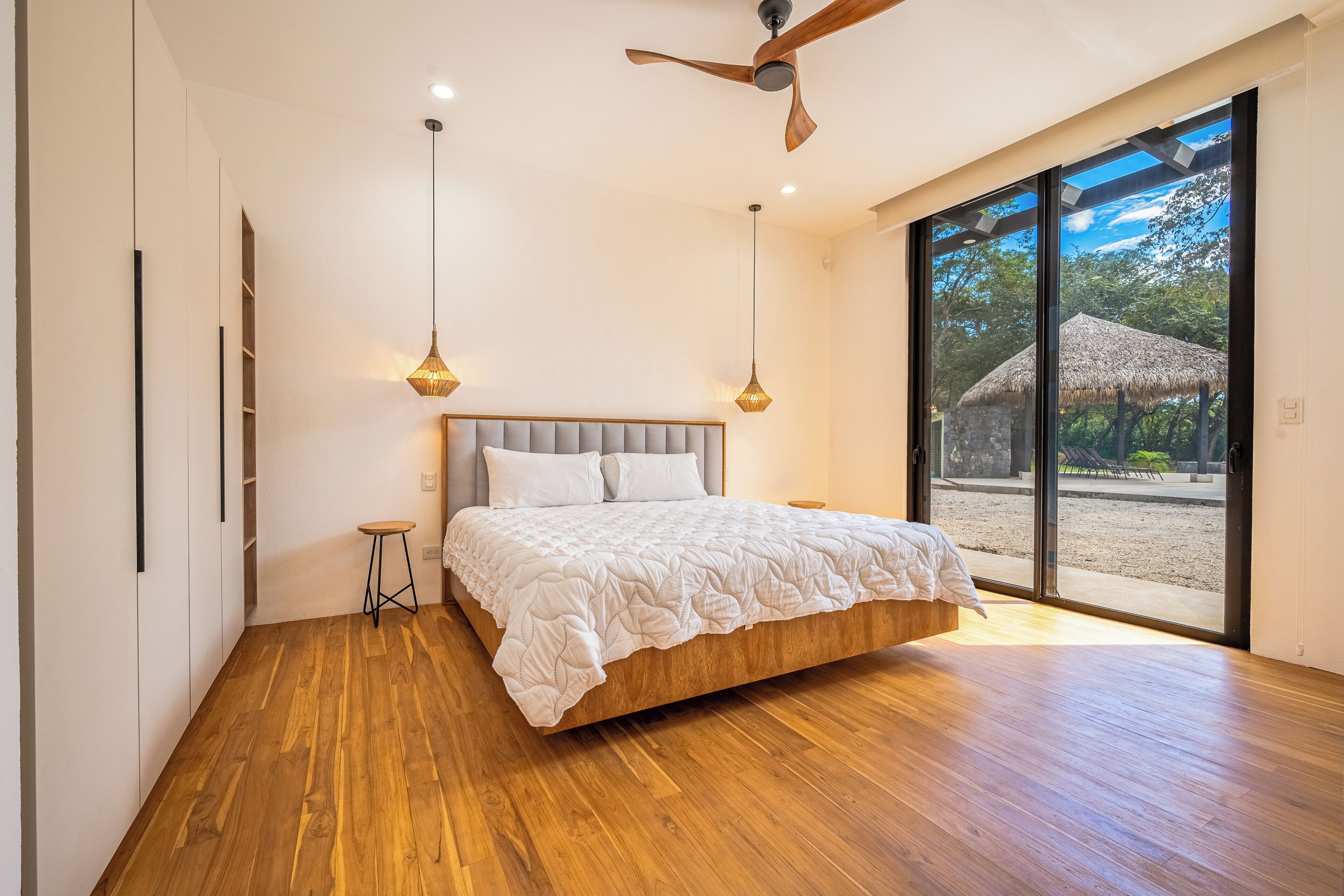 A modern bedroom with a large bed, wooden flooring, a ceiling fan, and a view of an outdoor area.