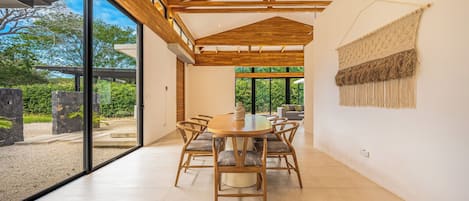 A spacious dining area with a wooden table and chairs, large windows, and a view of greenery outside.