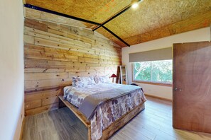 Ein Schlafzimmer mit Holzboden, einem Bett mit gemusterten Textilien, einem Fenster mit Blick auf Grünflächen und einer Tür.