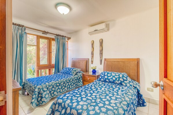A bedroom with two beds, a window with curtains, a bedside table, and a wall-mounted air conditioner. - Ocean Views: Bahia Pez Vela Resort Condo (Playa Ocotal)