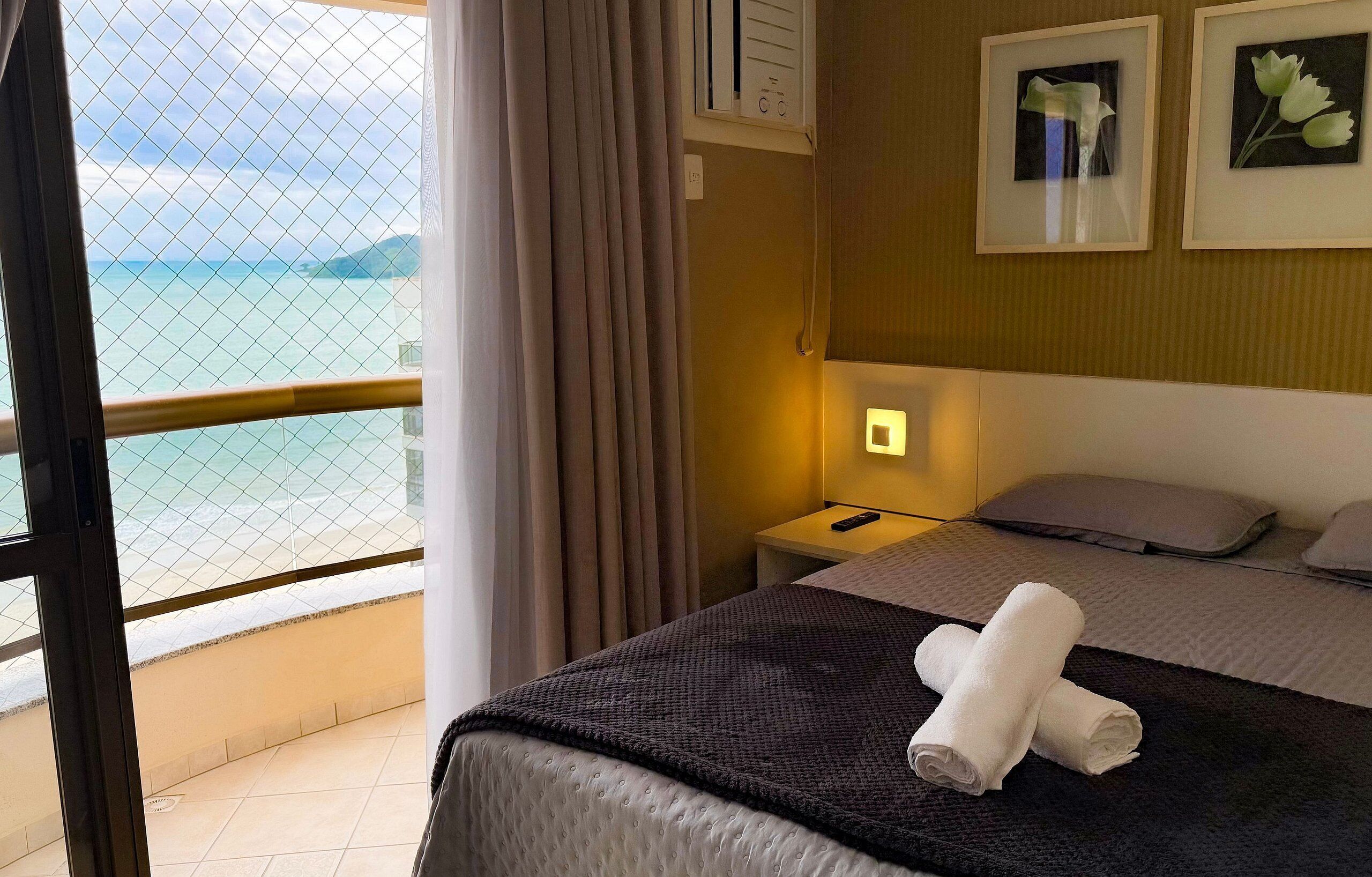 Una habitación de hotel con cama, mesita de noche, lámpara, ventana con cortinas y vista al mar.