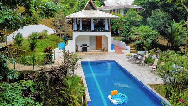 Piscina com área de descanso, cercada por vegetação exuberante e uma casa com telhado peculiar.