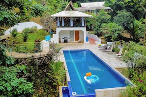 Piscina com área de descanso, cercada por vegetação exuberante e uma casa com telhado peculiar.