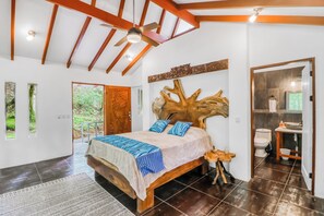 Une chambre avec un grand lit, un ventilateur de plafond, une tête de lit en bois et une vue sur l’extérieur par une porte.