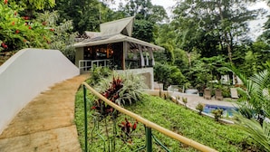 Um edifício moderno com telhado inclinado, rodeado por vegetação exuberante e área de piscina.