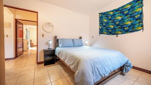 Quarto com cama, abajures, espelho fixado na parede e porta que dá para outro cômodo.