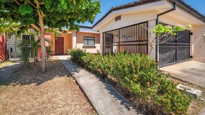 Edifício residencial com entrada por portão, pequena área de jardim com arbustos e uma árvore, e acesso por calçada.