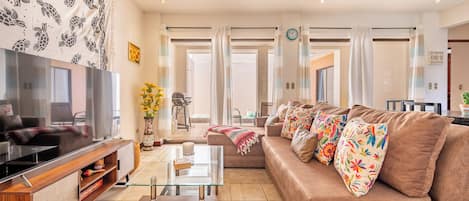 A modern living room with a flat-screen TV, a glass coffee table, a sofa with colorful pillows, and a patterned tapestry on the wall.