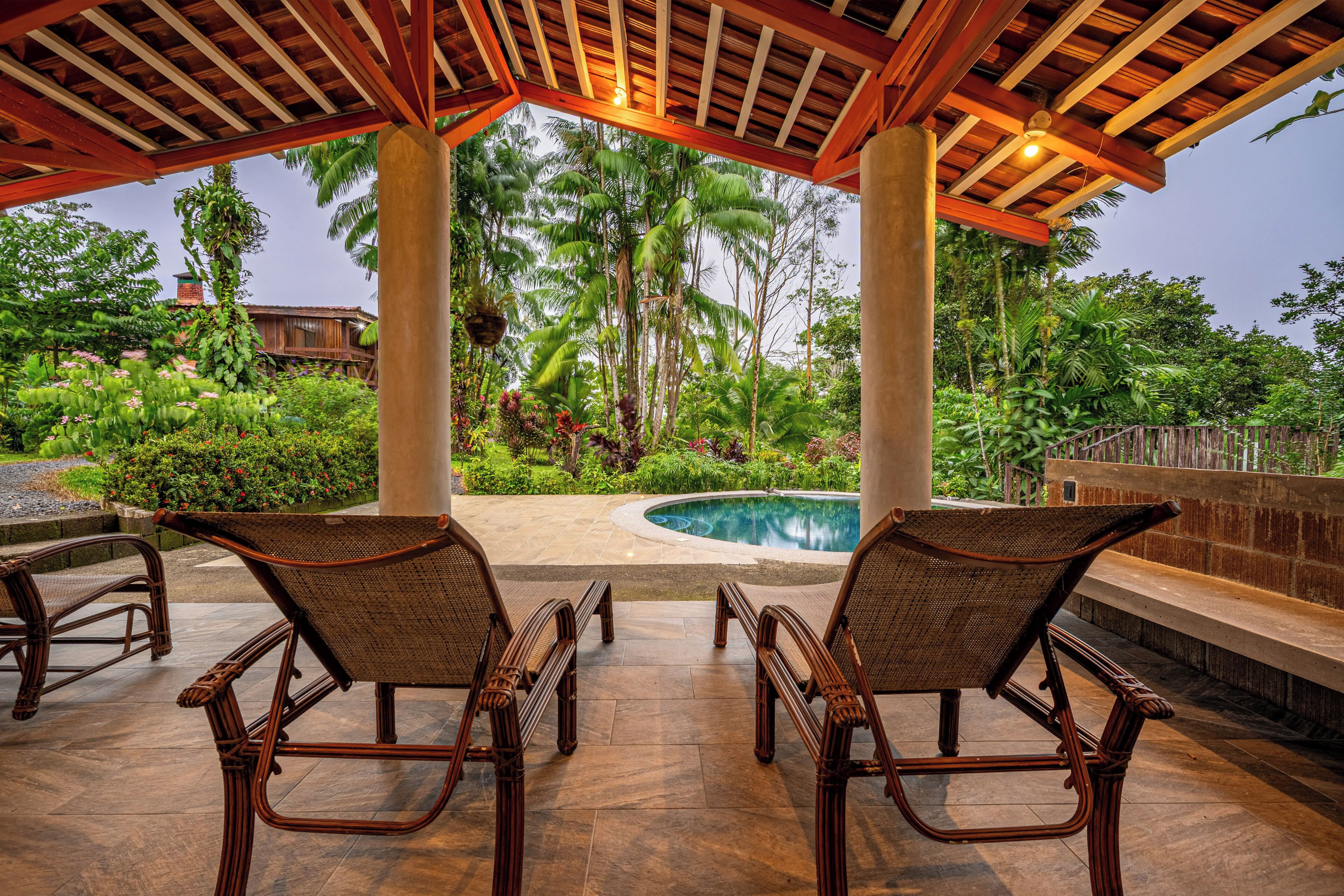 Un espacio al aire libre techado con piscina, sillones de descanso y vegetación tropical.