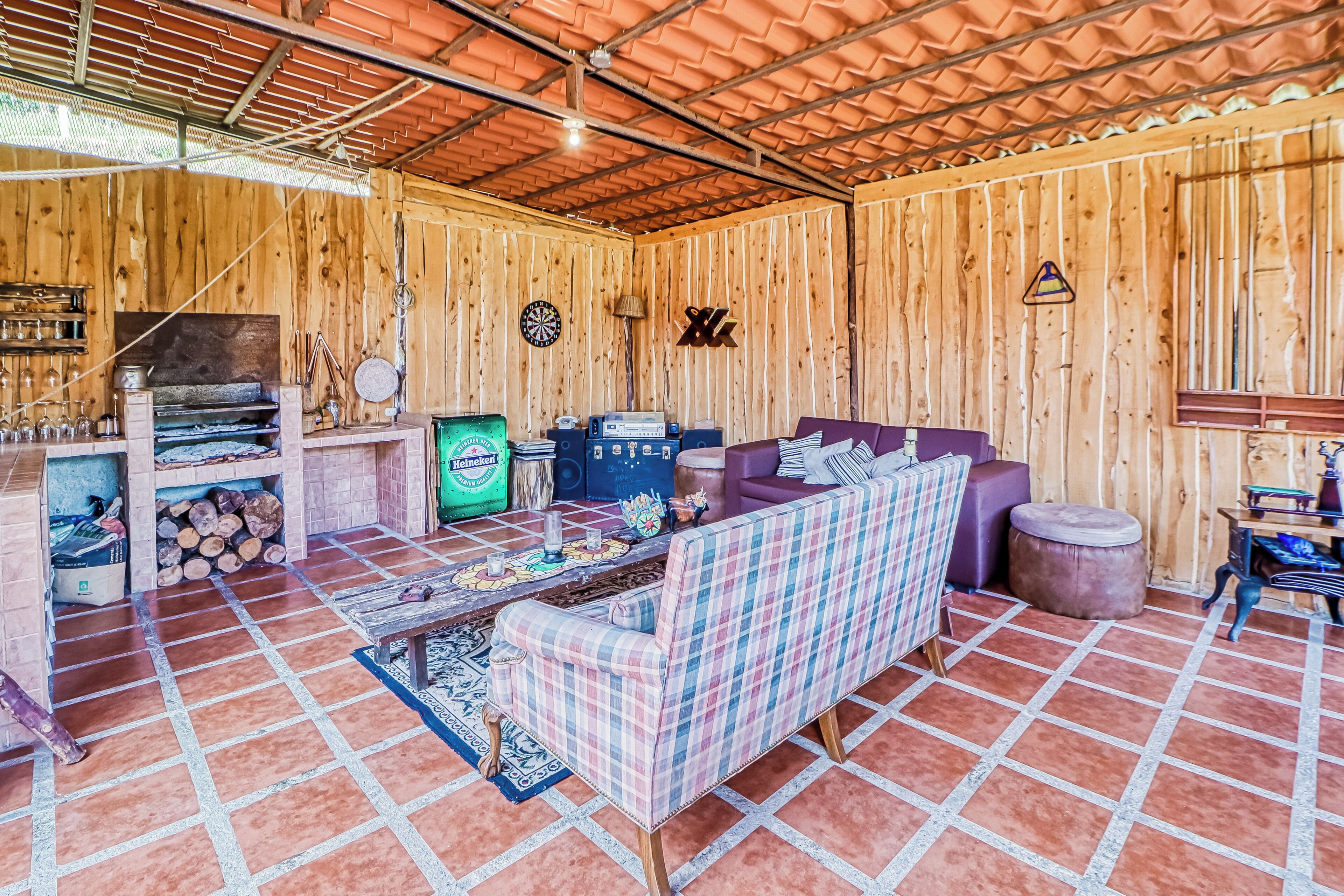 A rustic wooden interior with a checkered sofa, a table with a decorative tablecloth, and a wooden wall with a clock and a dartboard.