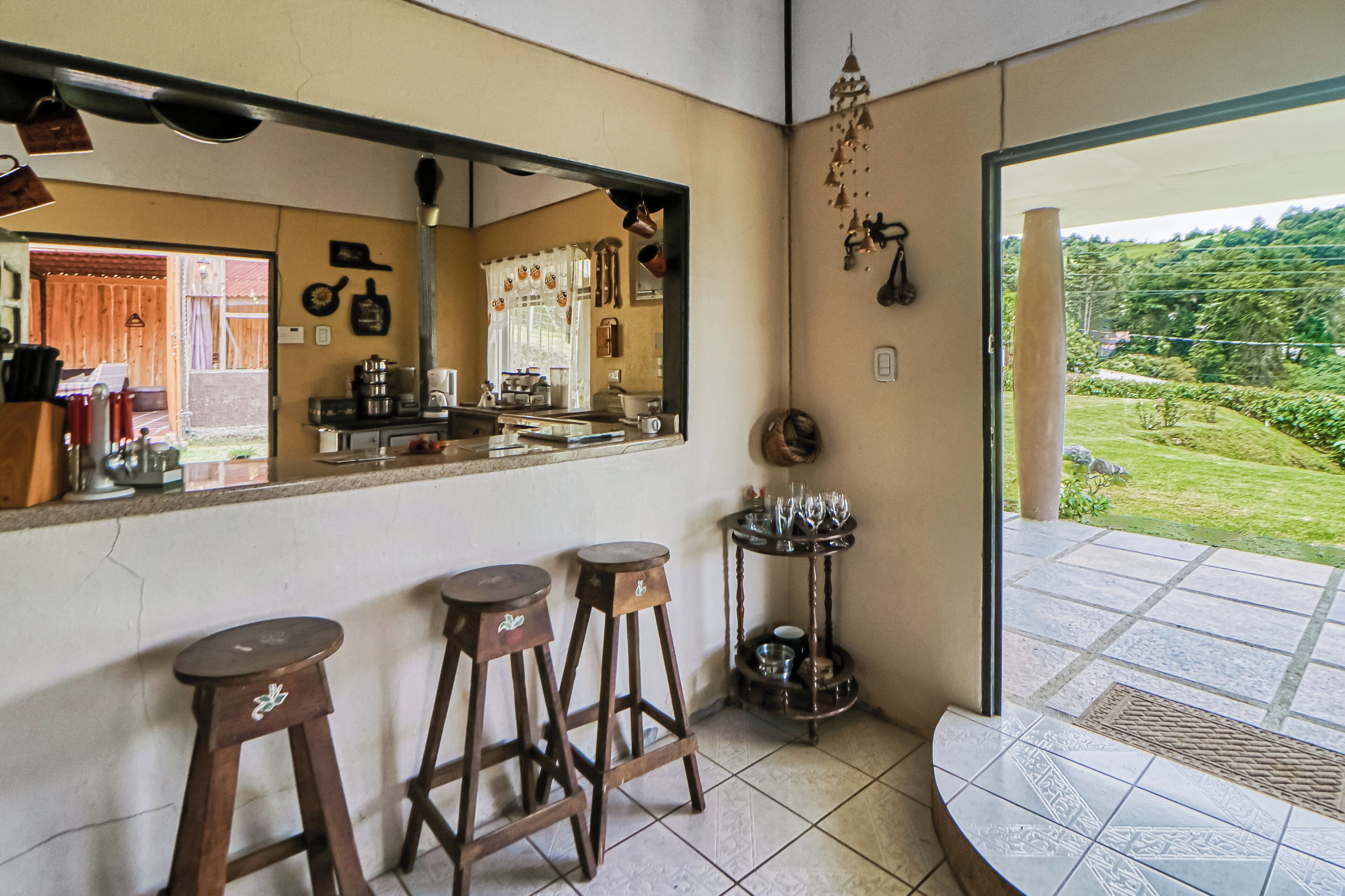 A kitchen with a large mirror, two stools, a coffee machine, and a view of a garden through an open door.