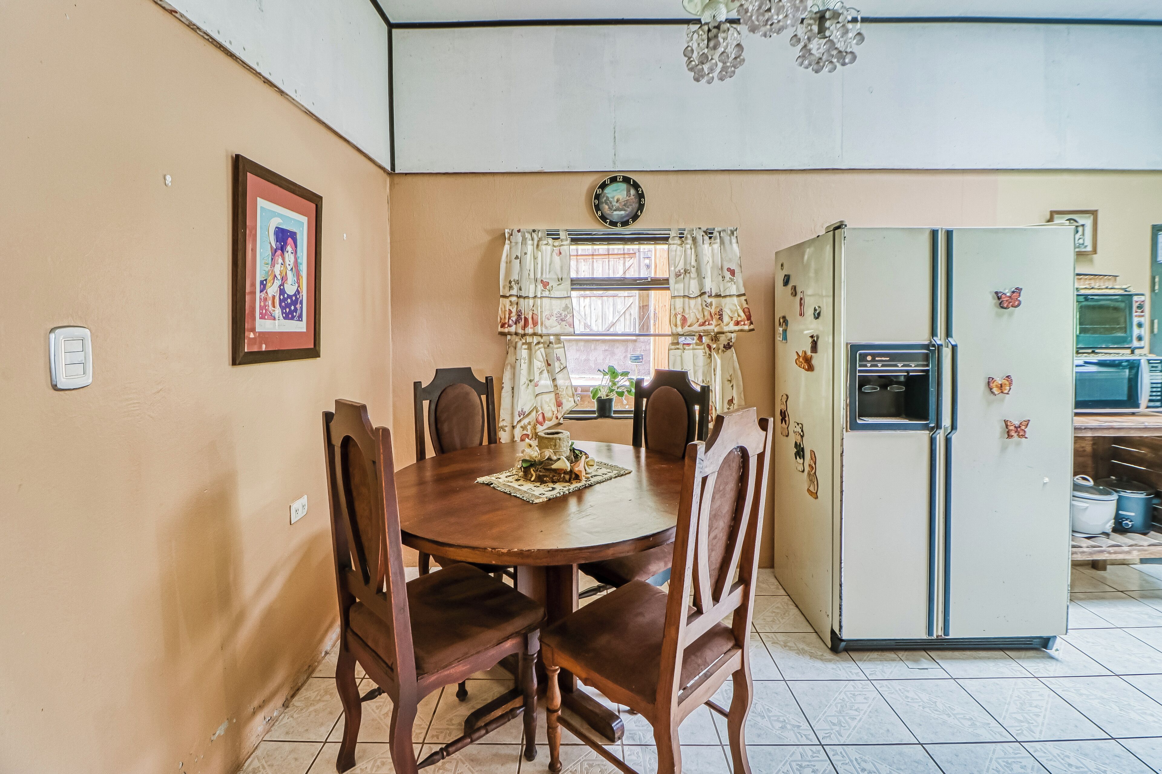 A dining area with a wooden table and chairs, a refrigerator with magnets, and a clock on the wall.