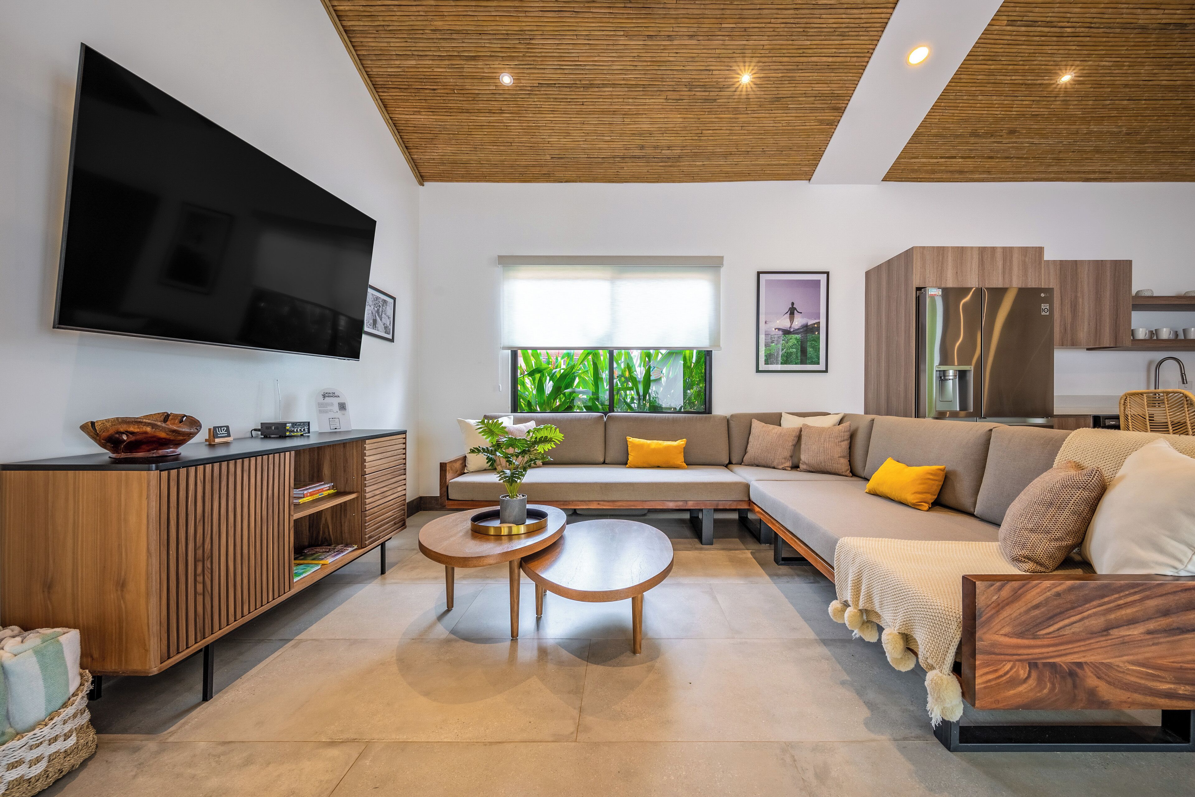 Un salon moderne avec un téléviseur à écran plat, un meuble en bois, un canapé avec des coussins, une table basse et une grande fenêtre.