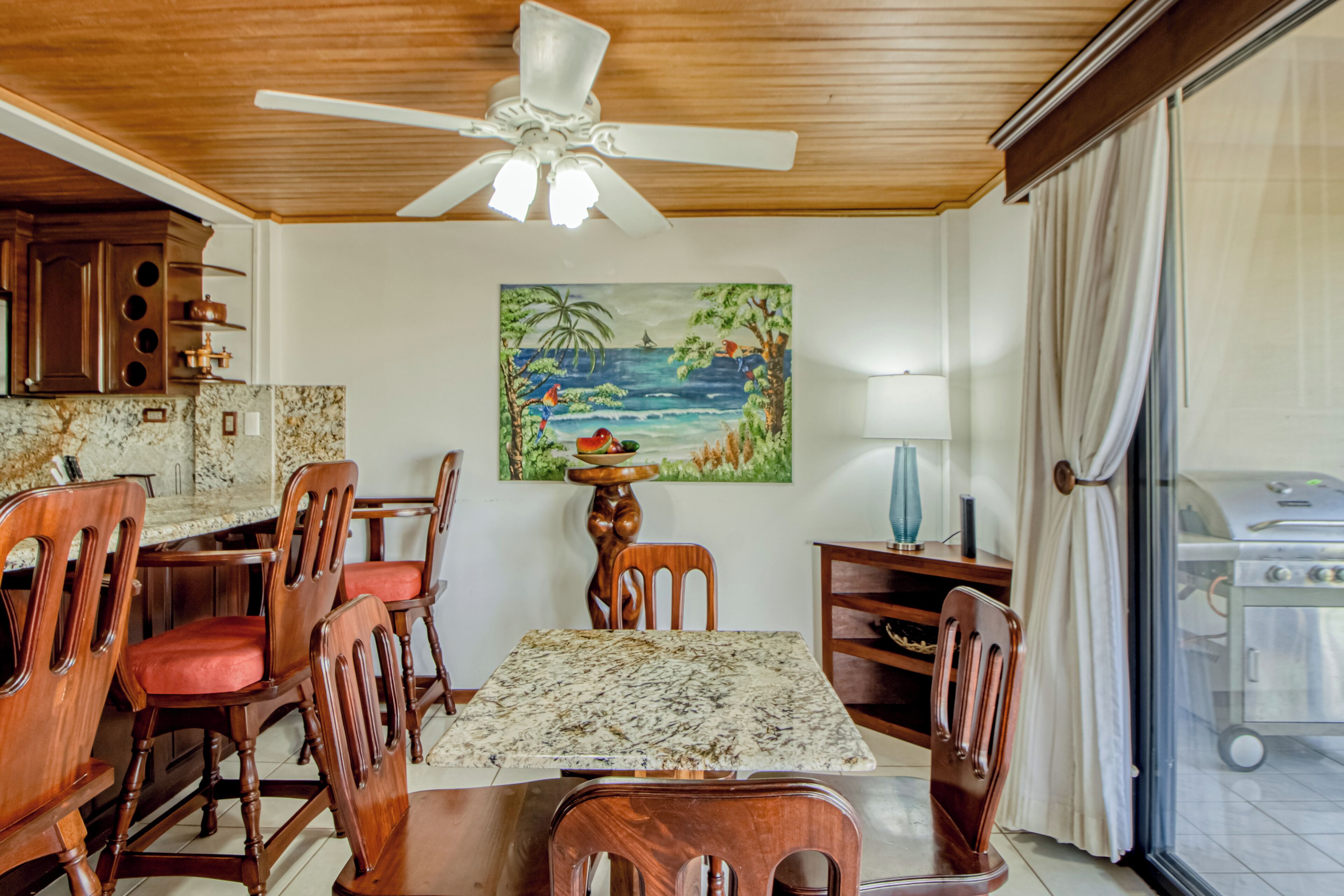 A dining area with a wooden table and chairs, a ceiling fan, a painting of a tropical scene, and a view through a glass door.
