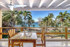 A wooden deck with a table and chairs, overlooking a beach with palm trees and the ocean. - Ideal 2BR Oceanfront 2nd-Floor | Balcony (Flamingo Beach)