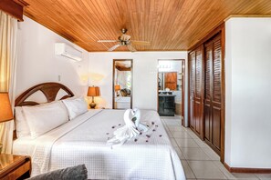 A hotel room with a bed, a ceiling fan, a nightstand, a lamp, a mirror, and a view of the bathroom. - Ideal 2BR Oceanfront 2nd-Floor | Balcony (Flamingo Beach)