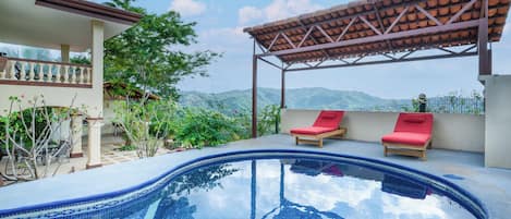 A swimming pool with two red lounge chairs under a pergola, surrounded by greenery and overlooking a mountainous landscape.
