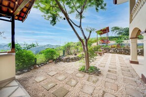 Eine Terrasse mit einem Steinweg, einer Pergola mit hängenden Lichtern, Blick auf die Berge und eine Steinmauer mit einer Bank.