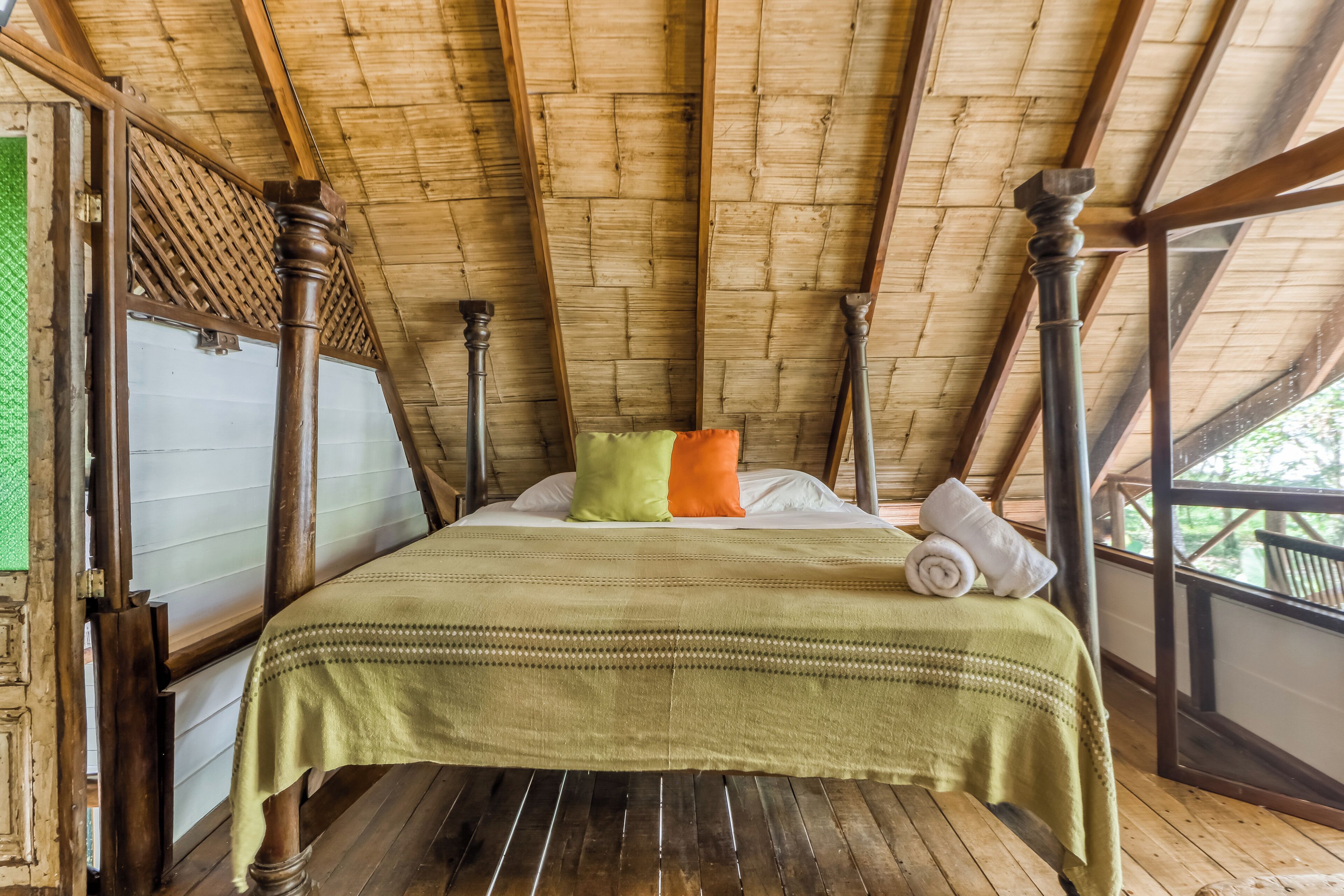 Ein Zimmer mit schrägen Wänden, Holzbalken, einem Bett mit grüner Bettdecke, Kissen und einem zusammengerollten Handtuch.