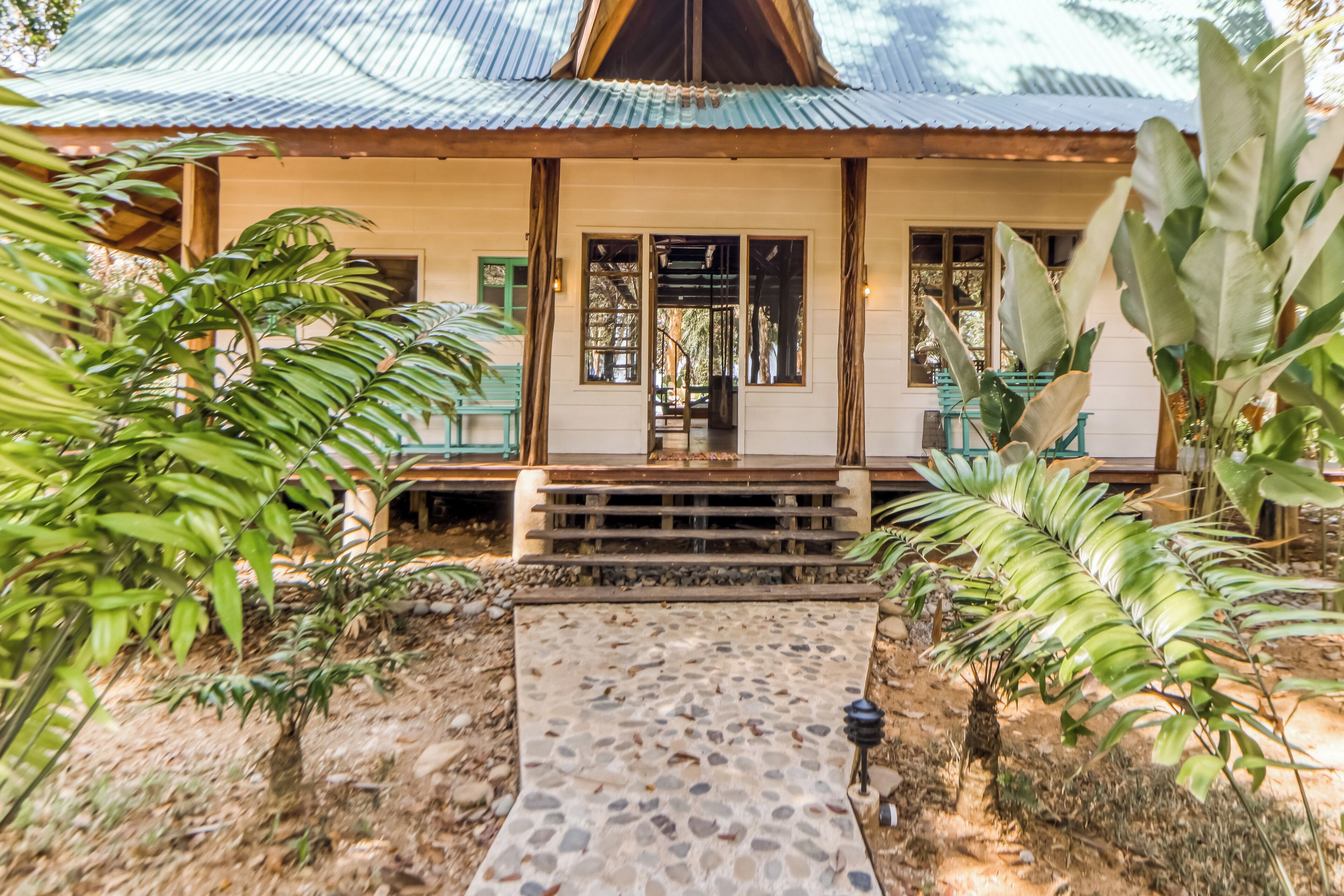 Une maison avec une véranda, un chemin de pierre menant à l’entrée et des plantes tropicales tout autour.