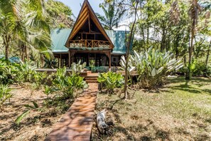 Une maison en bois avec un toit triangulaire, entourée d’une végétation tropicale et un sentier en bois menant à l’entrée.