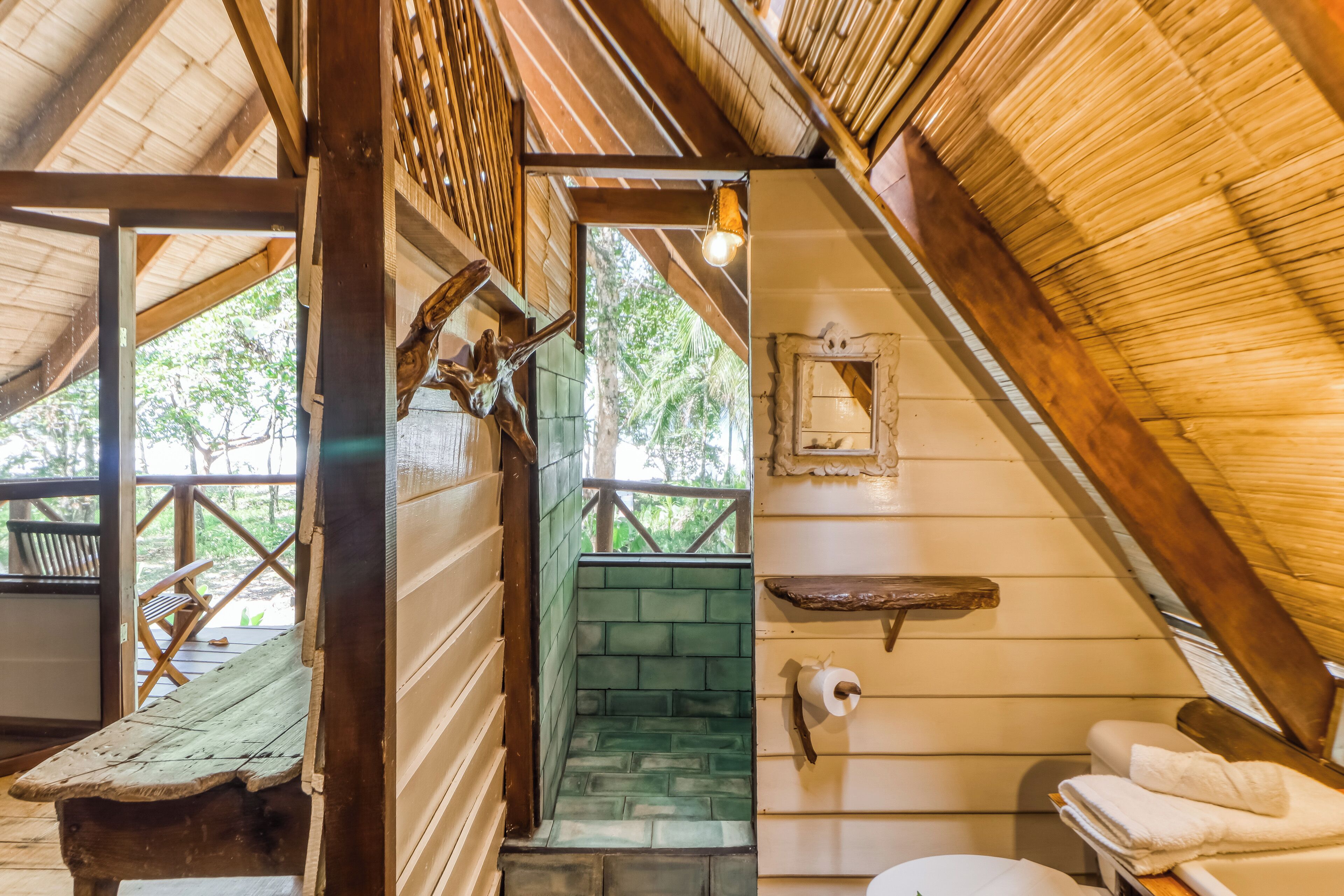 Un baño rústico con techo de madera, pared de azulejos verdes, banco de madera y un inodoro blanco.