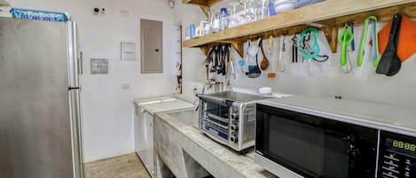 Een keuken met een roestvrijstalen koelkast, een magnetron en een elektrische oven. Boven het aanrecht staan planken met servies en bestek.