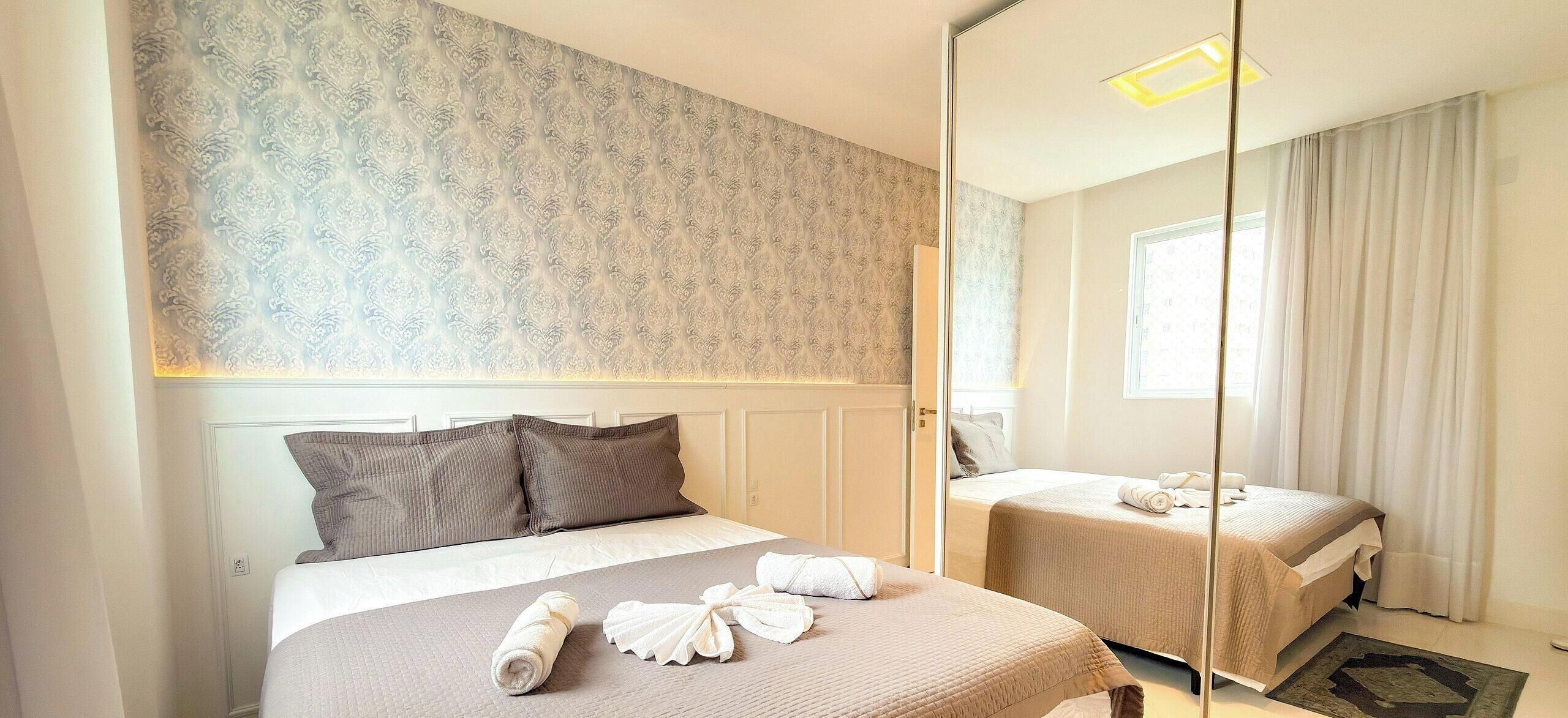 A hotel room with a large mirror, two beds, a patterned wallpaper, and a window with curtains.