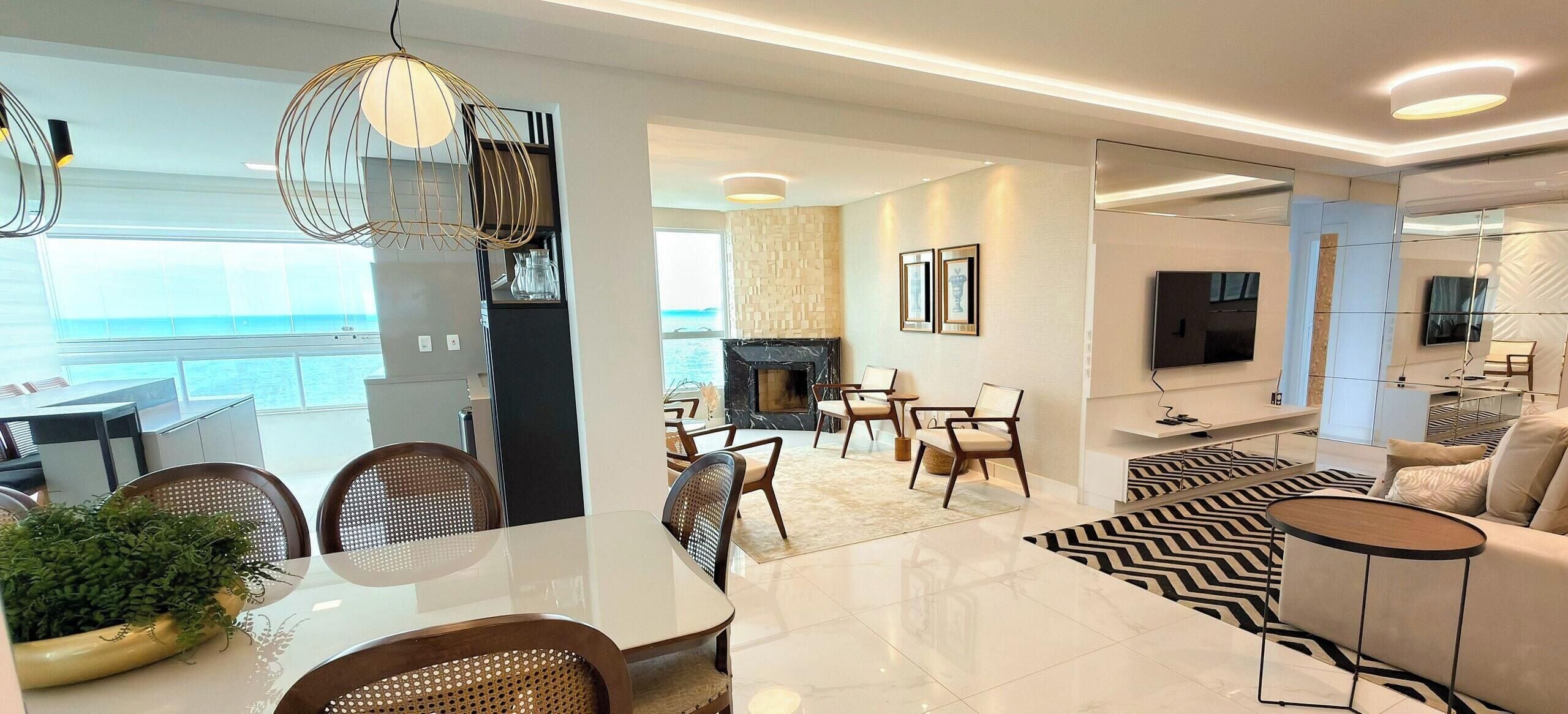 A modern living room with a fireplace, a dining area with wicker chairs, and a view of the sea.