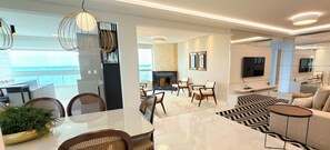 A modern living room with a fireplace, a dining area with wicker chairs, and a view of the sea. - ASA Cobertura Luxo 2QT Meia Praia Hidro (Itapema)