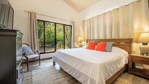 Quarto com cama grande, uma cadeira, mesa de cabeceira e janela com cortinas.