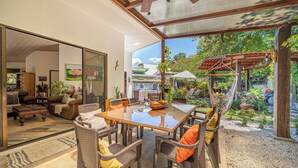 Uma sala de estar moderna com parede de vidro, área de jantar com mesa e cadeiras de madeira, e vista para um jardim com pérgula.