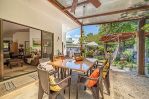 A modern living room with a glass wall, a dining area with a wooden table and chairs, and a view of a garden with a pergola. - Pool & Large Patio: Playa Carrillo 3BR Family Home (Playa Carrillo)