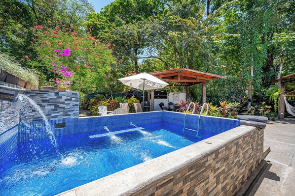 A swimming pool with a stone wall and tiled floor, surrounded by lush greenery and a wooden pergola. - Pool & Large Patio: Playa Carrillo 3BR Family Home (Playa Carrillo)