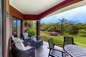 A covered outdoor seating area with wicker furniture and a glass table, overlooking a lush garden with various plants and a palm tree. - Playa Conchal Resort Condo w/ Ocean View (Playa Conchal)