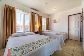 A hotel room with two beds, a window with curtains, a wall-mounted air conditioner, a door, and a small table with a lamp. - Playas del Coco Condo w/ Pool: Walk to Beach! (Playas del Coco)
