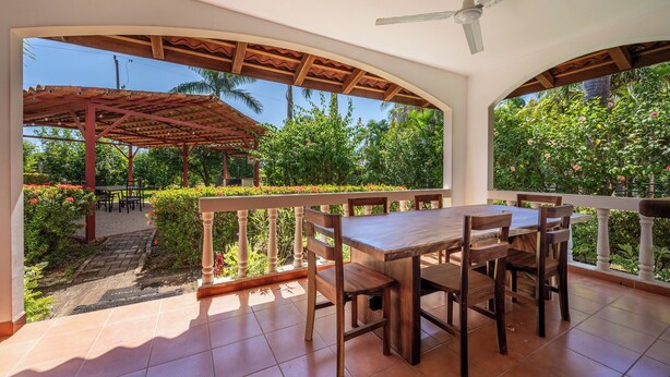 Área de refeições ao ar livre coberta, com mesa e cadeiras, ventilador de teto e vista para um jardim com diversas plantas e flores.