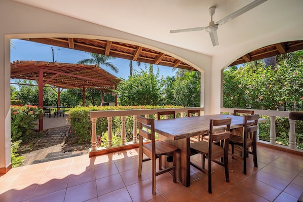 A covered outdoor dining area with a table and chairs, a ceiling fan, and a view of a garden with various plants and flowers. - Santa Cruz Group Getaway w/ Pool: Walk to Beach (Junquillal)