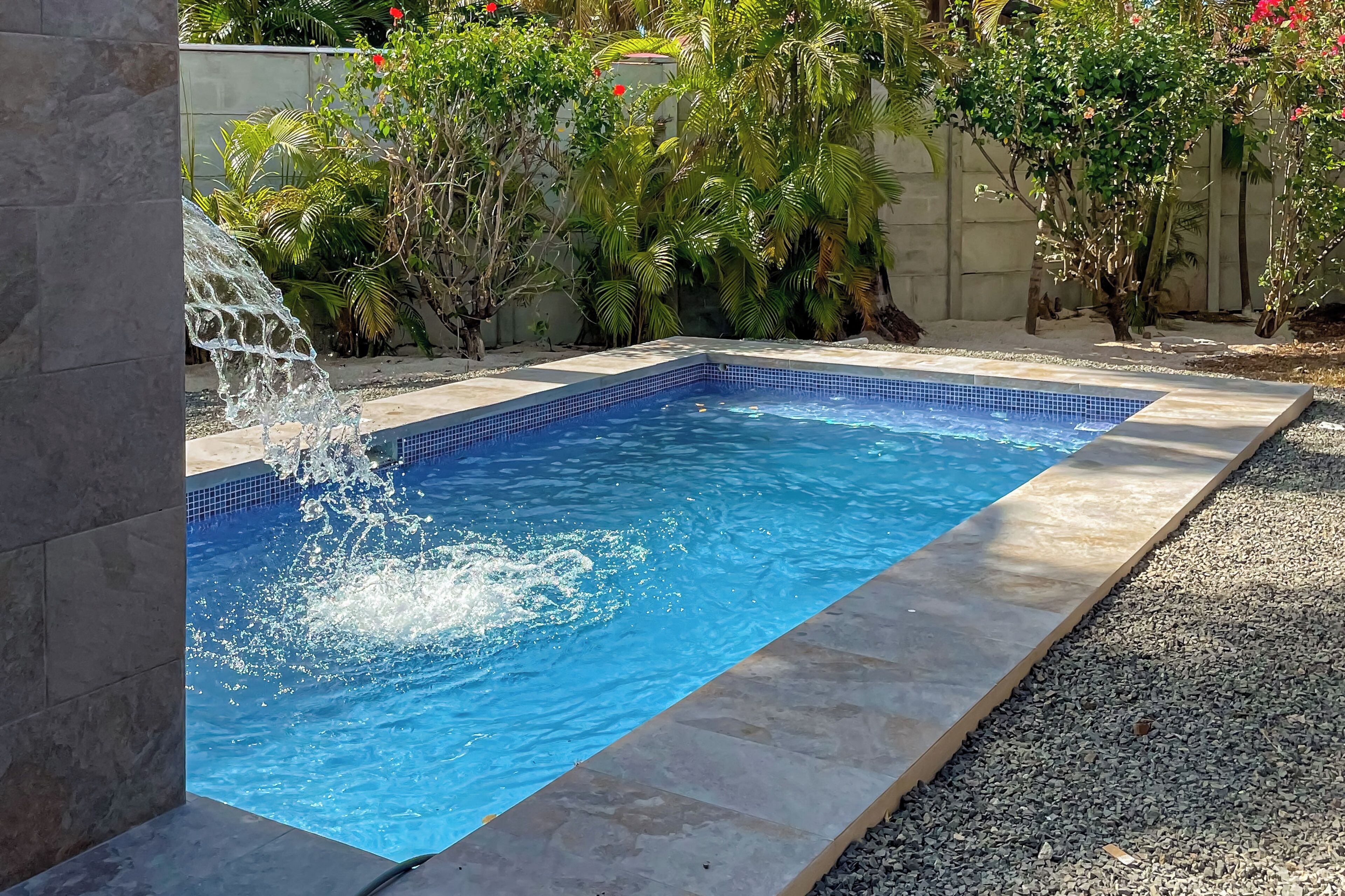 A rectangular swimming pool with a tiled edge, surrounded by a gravel area and various plants.