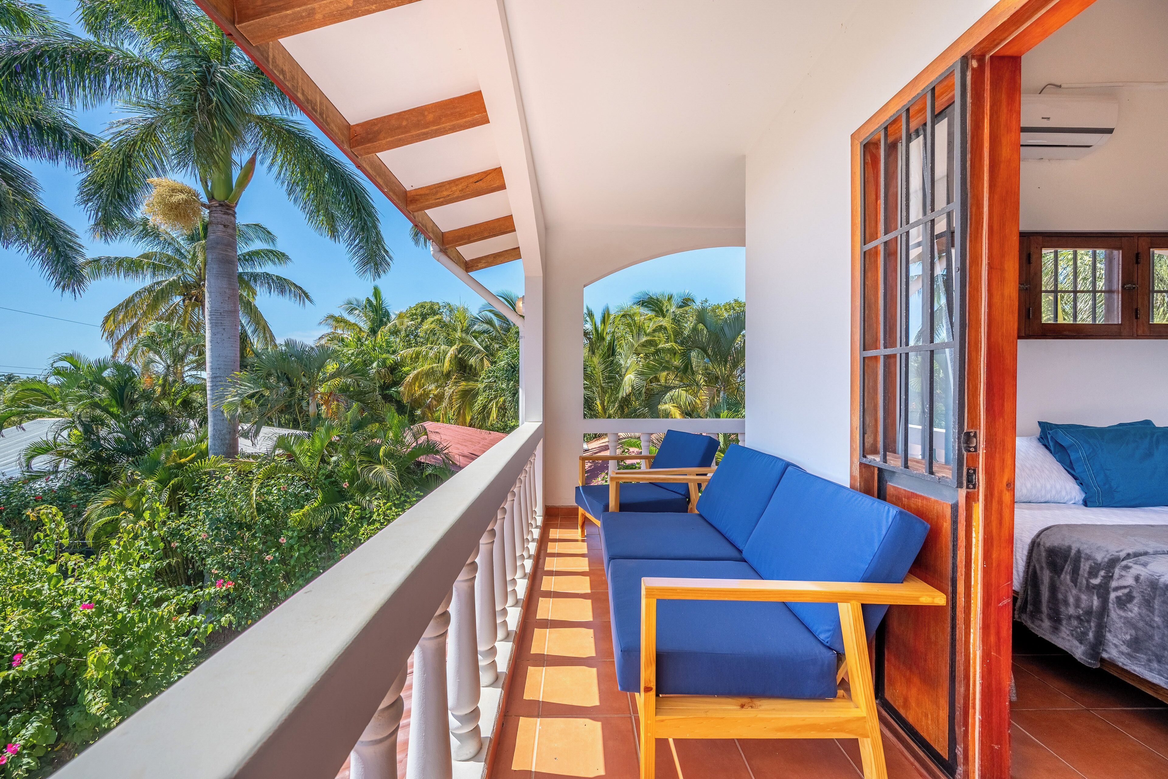 Een balkon met een bank en stoelen, een bed met blauw beddengoed, en uitzicht op palmbomen en groen.