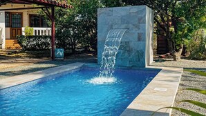 Piscina com cascata, cercada por um muro de pedra e uma área coberta com escadas que levam a uma porta.