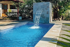 A swimming pool with a waterfall feature, surrounded by a stone wall and a covered area with steps leading to a door. - Santa Cruz Group Getaway w/ Pool: Walk to Beach (Junquillal)