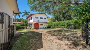 Uma casa de dois andares com uma porta vermelha, cercada por um gramado bem cuidado e várias árvores.