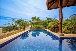 A clear blue swimming pool with tiled edges, surrounded by lush greenery and a wooden pergola structure. - Luxe Ocean-View Hideaway in Carrillo (Playa Carrillo)