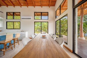 A spacious dining area with a long wooden table, multiple chairs, and large windows offering views of greenery. - Luxe Ocean-View Hideaway in Carrillo (Playa Carrillo)